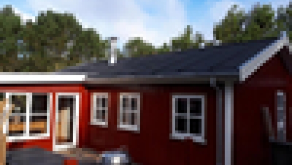 Udvendigt billede af et rødt hus med nyt tag, omgivet af træer og blå himmel.